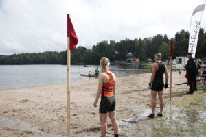 Kuvassa hiekkarannalla kaksi urheilijja valmiina juoksemaan vesirajassa olevien veneiden luokse ja soutamaan.
