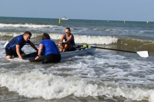 Rannan tuntumassa kaksi henkilöä auttamassa coastal-soutajaa aaltojen murtuessa valkoisena.